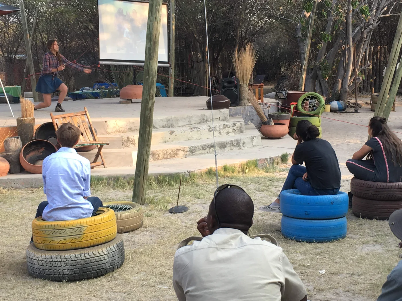 A student performs on an outdoor community stage while classmates and local hosts watch from seats made of painted tires